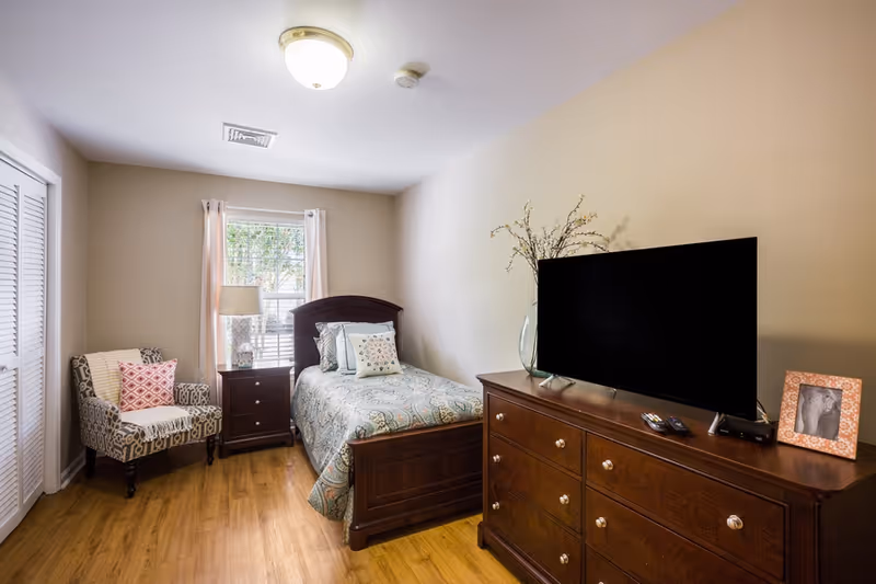 A furnished single bedroom featuring a twin bed, wooden dresser with a TV, nightstand, and an upholstered chair by a window.
