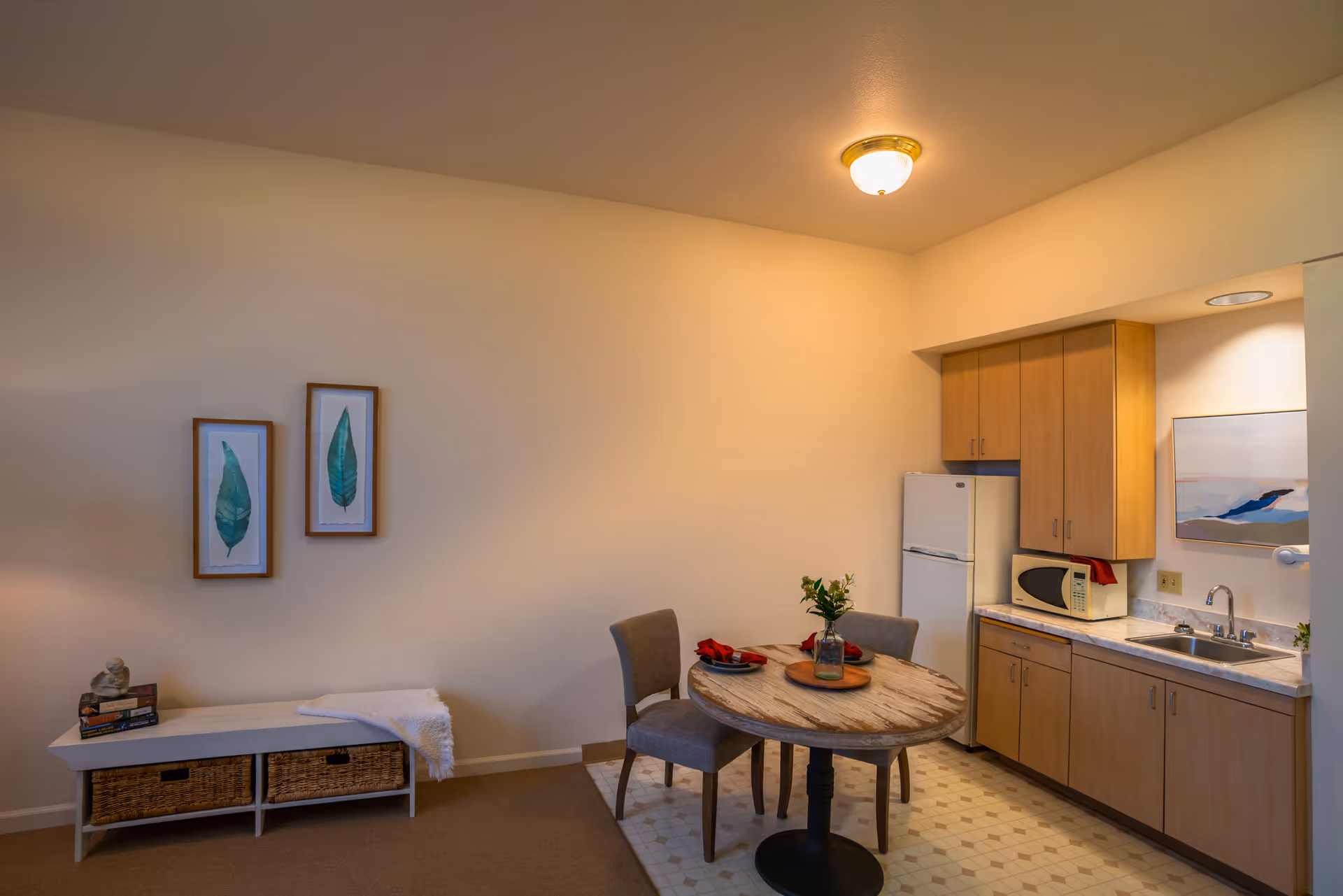 Small studio-style interior with a kitchenette, round dining table and two chairs, and wall art above a storage bench.