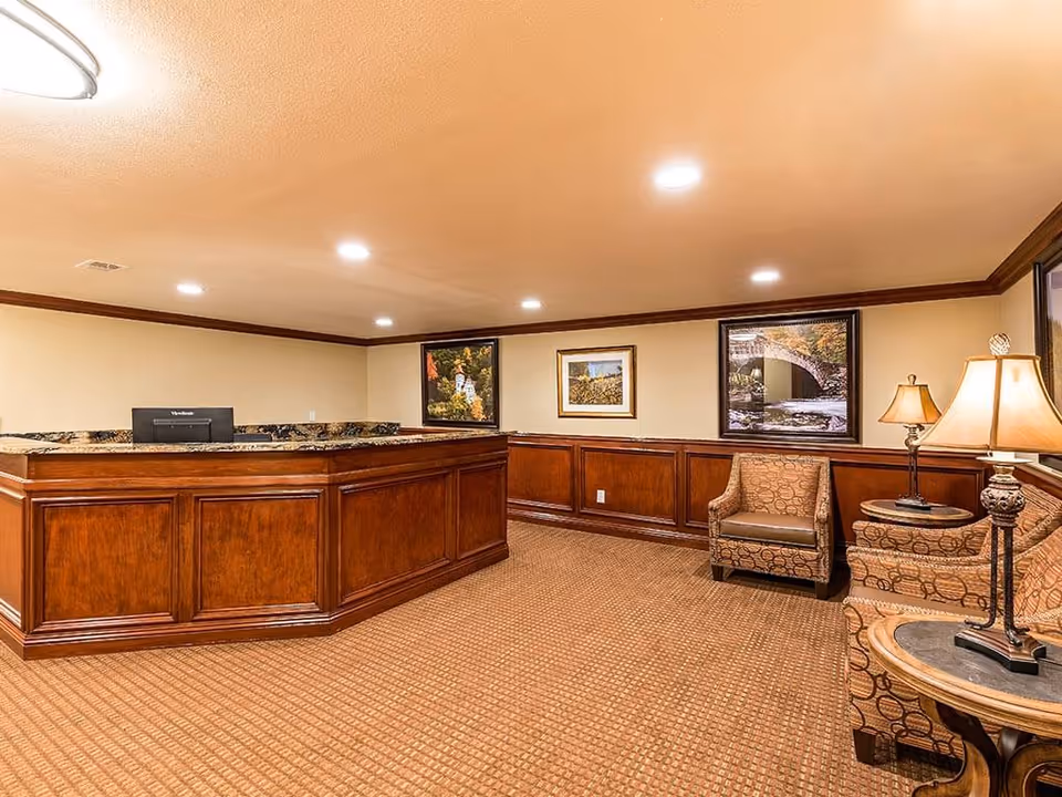 Reception area with a wooden front desk, patterned carpet, two upholstered chairs, a side table with a lamp, and framed landscape pictures on the walls.