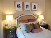 A neatly made bed with multiple pillows including a bolster pillow, two bedside tables each with a lamp, and three framed black and white photos hanging on the wall above the headboard.