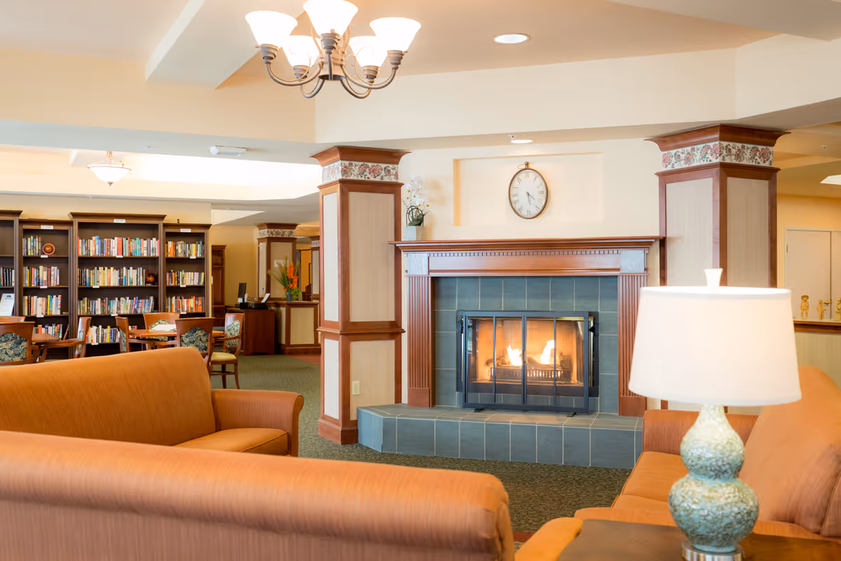 A cozy living room area in a senior living facility featuring two orange sofas facing a lit fireplace with a clock above it. There is a green ceramic table lamp on a side table next to one sofa. In the background, there is a bookshelf filled with books and several chairs and tables for reading or socializing. The room has warm lighting and decorative columns with floral trim.