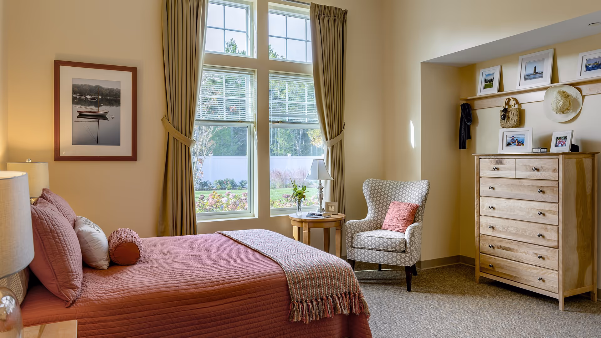 A cozy bedroom with a bed covered in a rust-colored quilt and pillows, a wooden nightstand with a lamp, a round wooden side table with a lamp and a small vase of flowers, a patterned armchair with a pink cushion, a wooden dresser with framed photos and a hat hanging above it, and large windows with beige curtains letting in natural light.