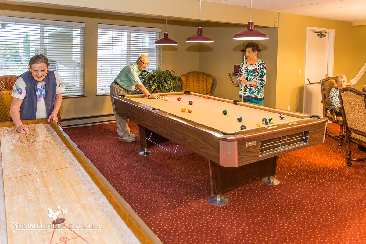 Four elderly individuals in a recreational room at Summerfield Estates Retirement Community. One woman is playing shuffleboard, a man is preparing to take a shot on a pool table, another woman is holding a pool cue, and a fourth woman is seated in a chair watching. The room has large windows with blinds, red carpet, and yellow walls.