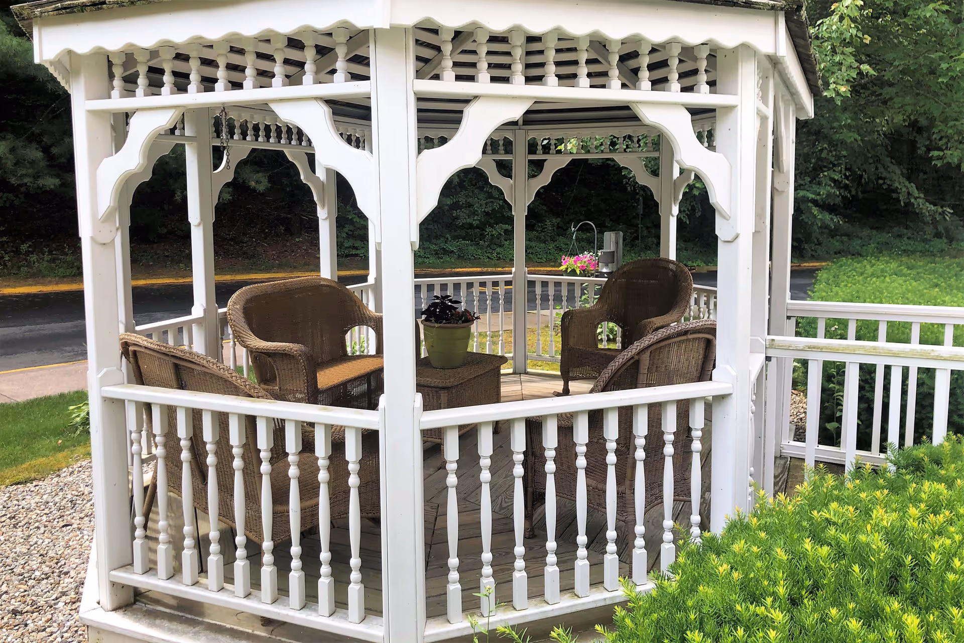 White wooden gazebo with decorative railings and roof trim, containing four wicker chairs and a small table with a potted plant, surrounded by greenery and a paved path nearby.