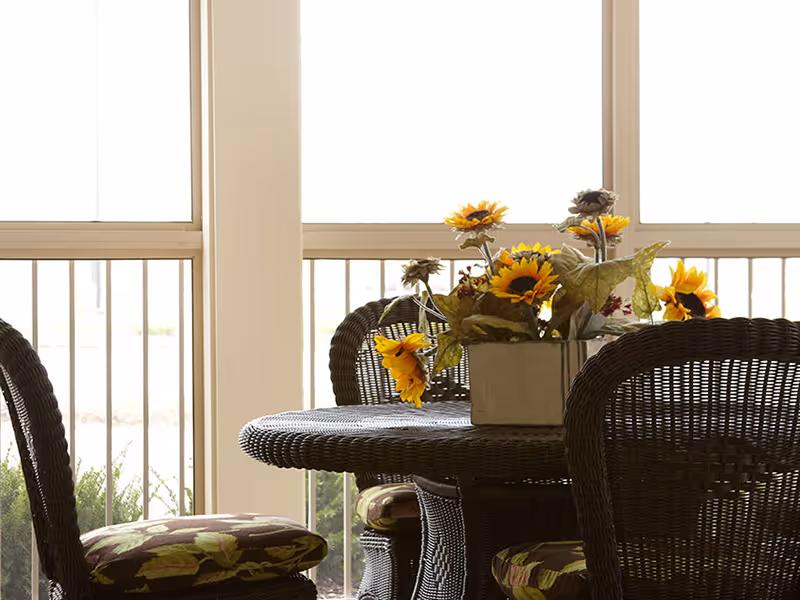 Wicker dining table and chairs with a sunflower centerpiece in front of large windows.