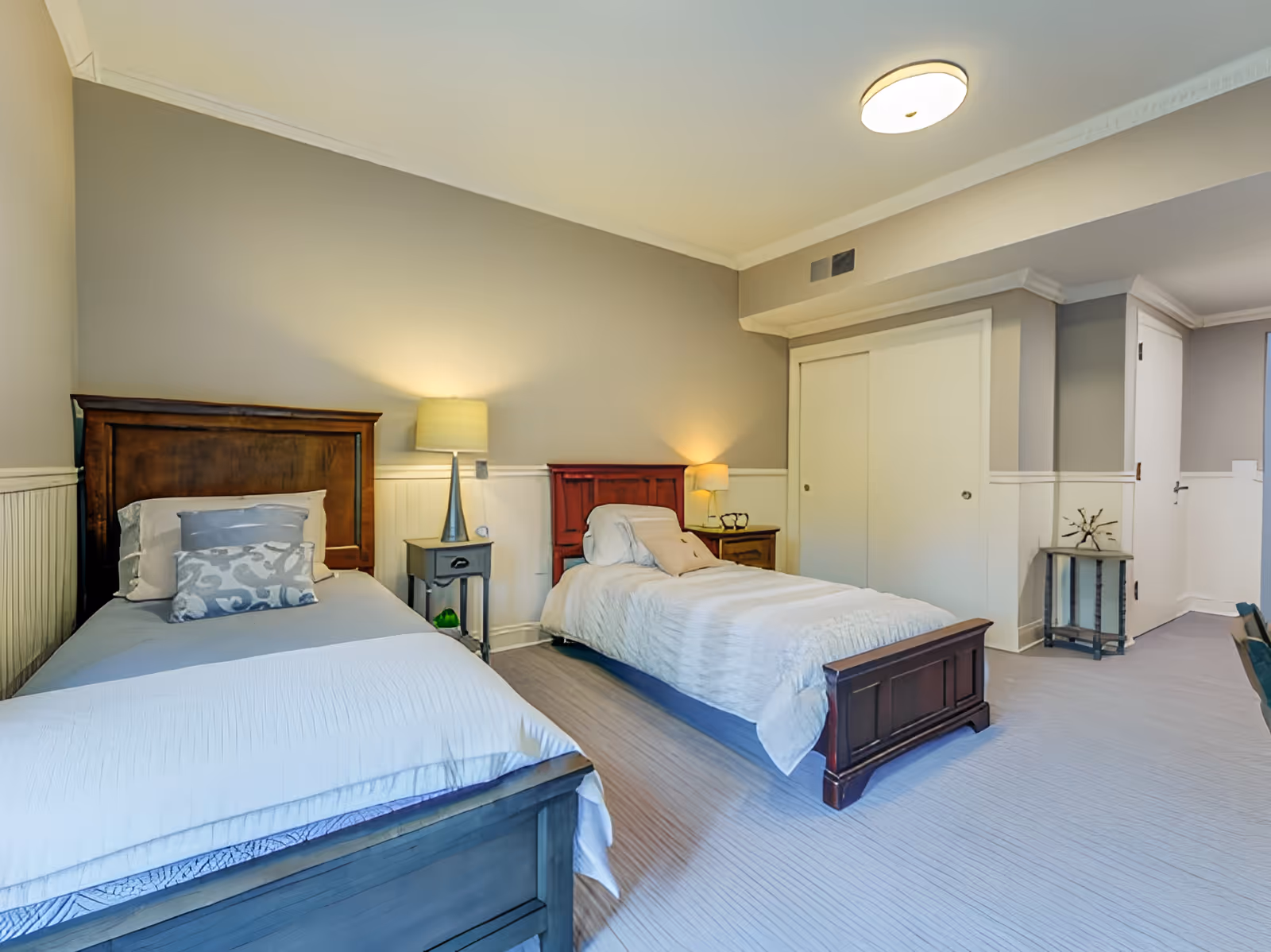 A bright and clean bedroom with two single beds, each with wooden headboards and white bedding. Between the beds are two nightstands with lamps. The walls are painted light gray with white wainscoting, and there is a closet with sliding white doors. The room has a modern ceiling light and light-colored flooring.
