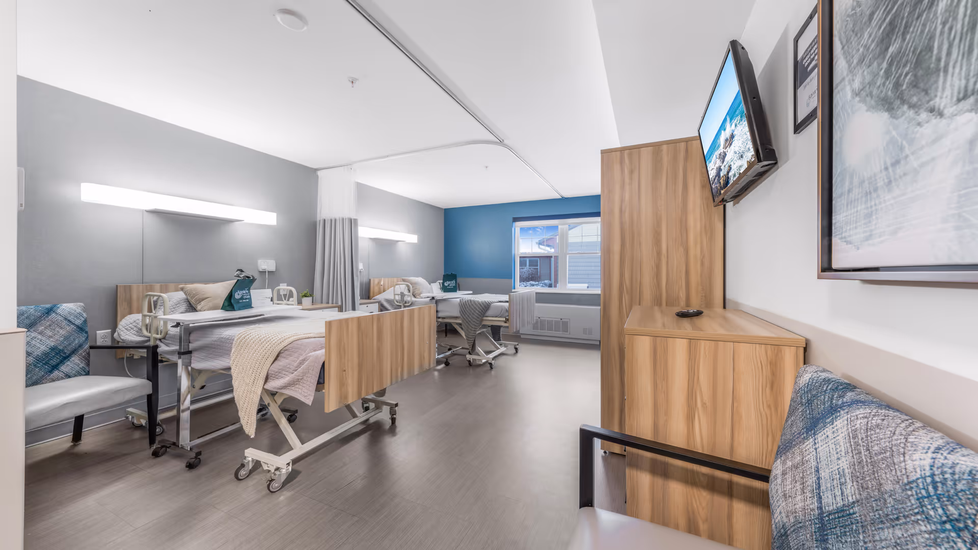 Bright shared rehabilitation room with two adjustable hospital beds, seating, a wall-mounted TV, and wooden cabinetry.
