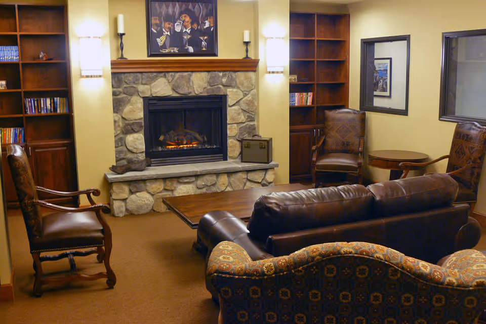 Cozy living room with a stone fireplace, leather sofa, patterned armchair, wooden coffee table, two wooden armchairs, built-in bookshelves, wall sconces, and framed artwork above the fireplace.