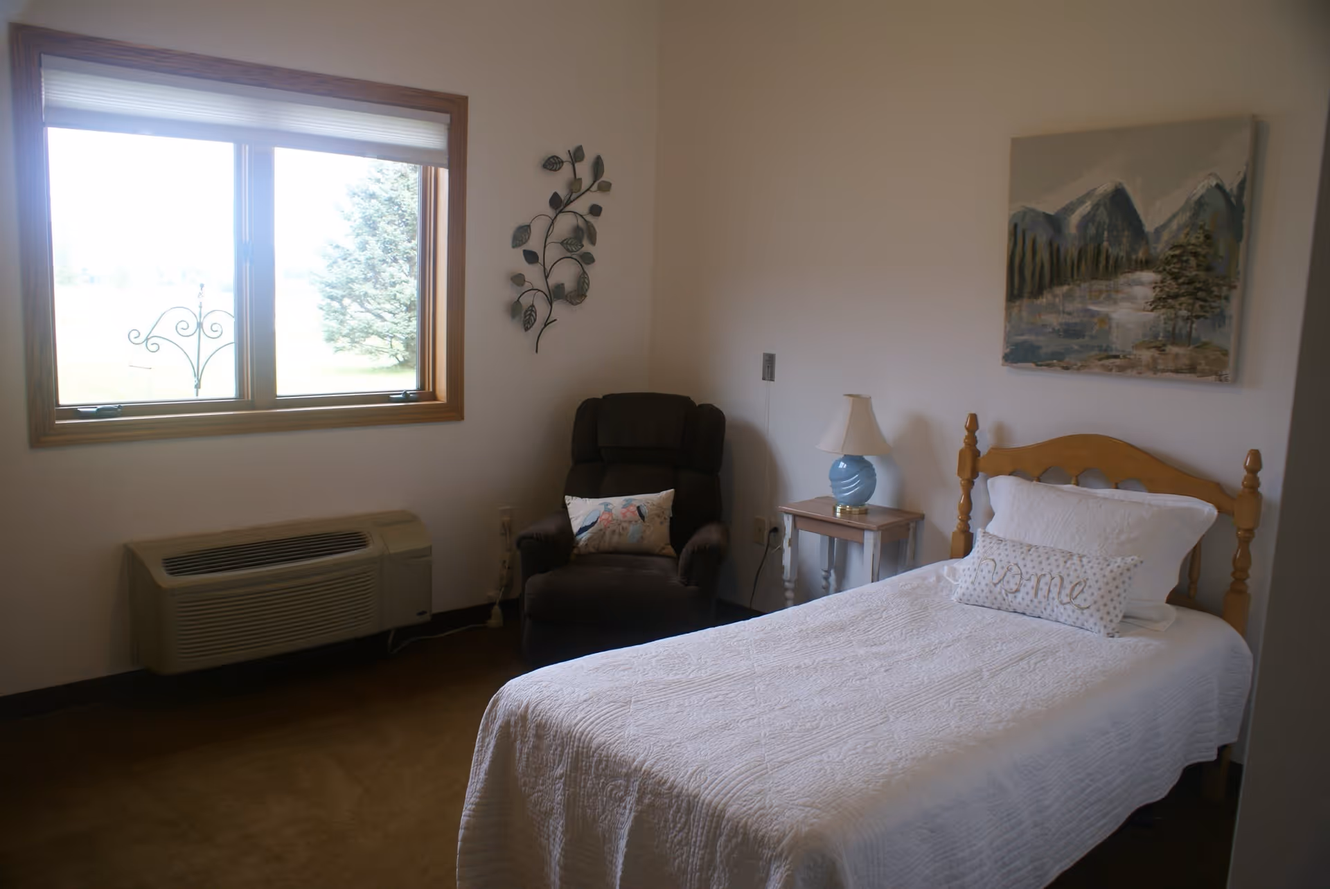 A simple bedroom with a single wooden bed covered in a white quilt and pillows, including one with the word 'Home'. Next to the bed is a small wooden nightstand with a blue lamp. A dark upholstered armchair with a decorative pillow is positioned near a window with a view of a tree outside. The walls are white with a metal leaf wall decoration and a landscape painting of mountains and trees.