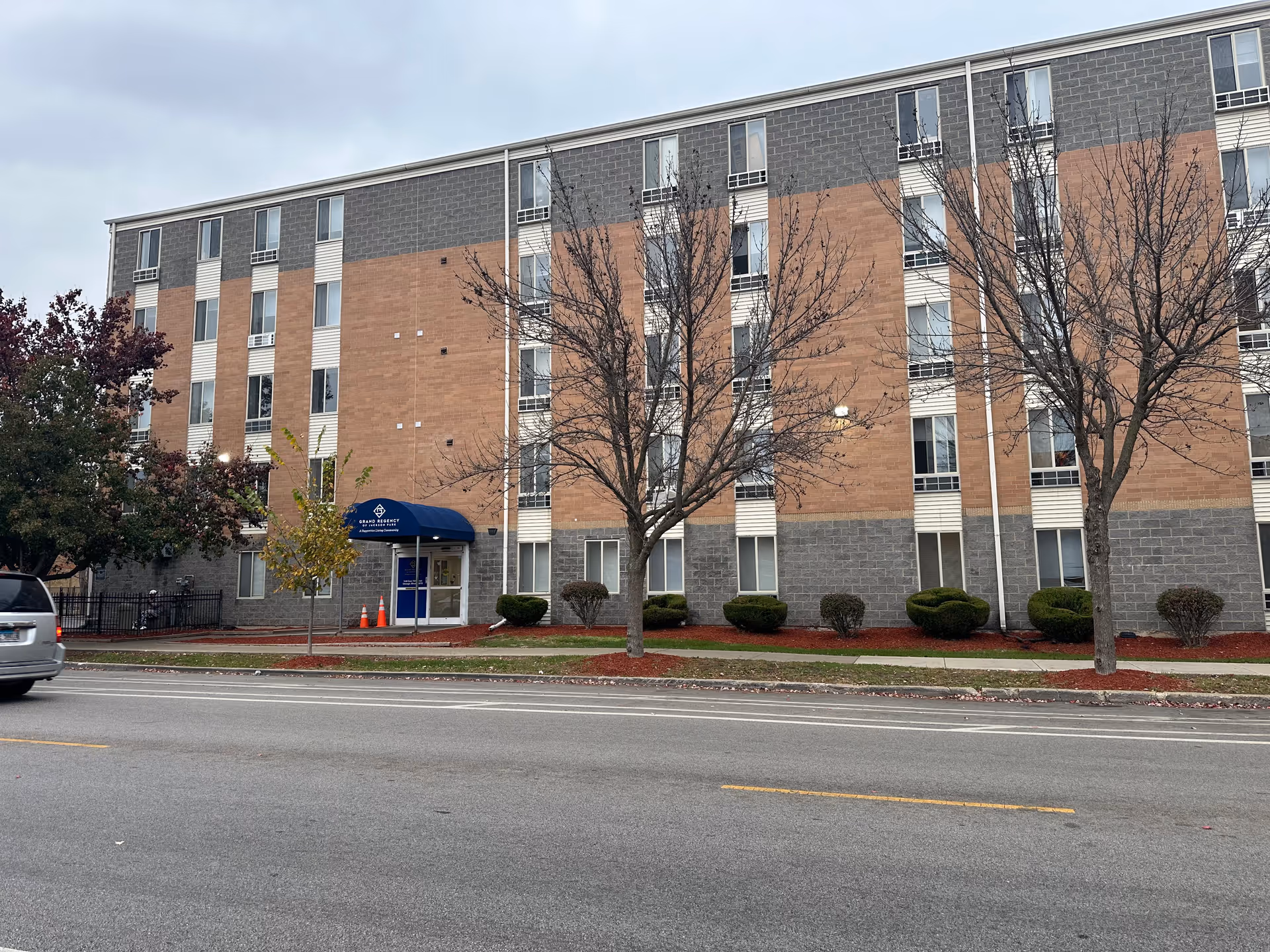 Front exterior of a multi-story brick supportive living building with a blue entrance canopy and trees along the sidewalk.
