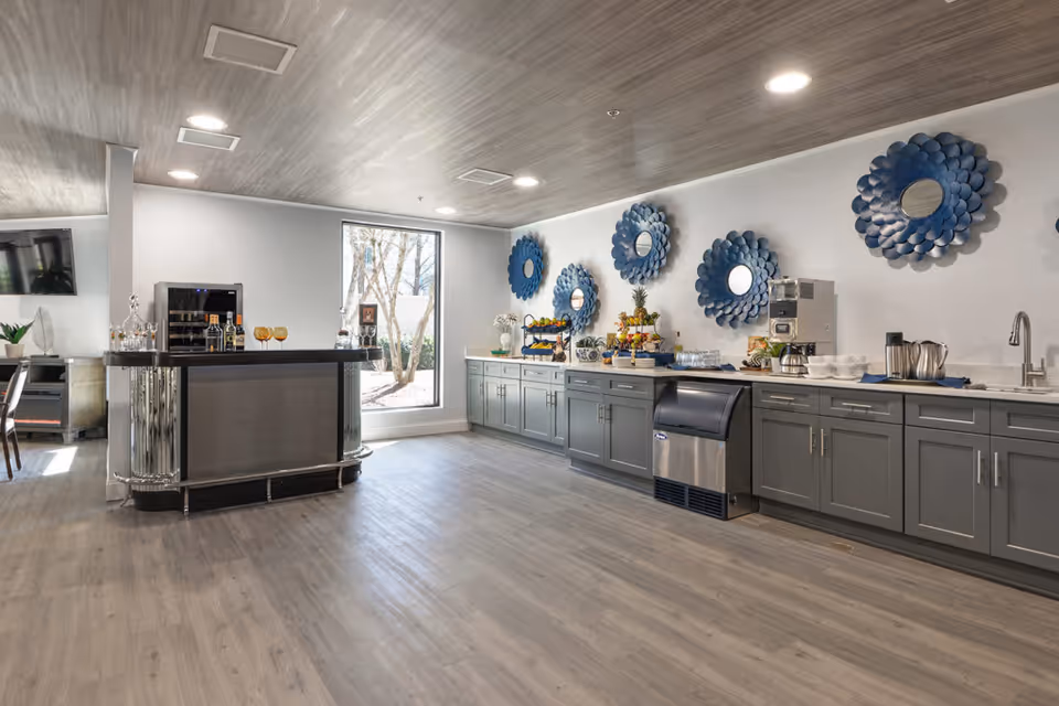 A spacious interior room with gray wooden flooring and ceiling, featuring a long counter with gray cabinets and various refreshments including fruit, coffee, and glasses. Blue decorative circular wall mirrors are mounted above the counter. A small bar area with bottles and glasses is on the left side near a large window letting in natural light.