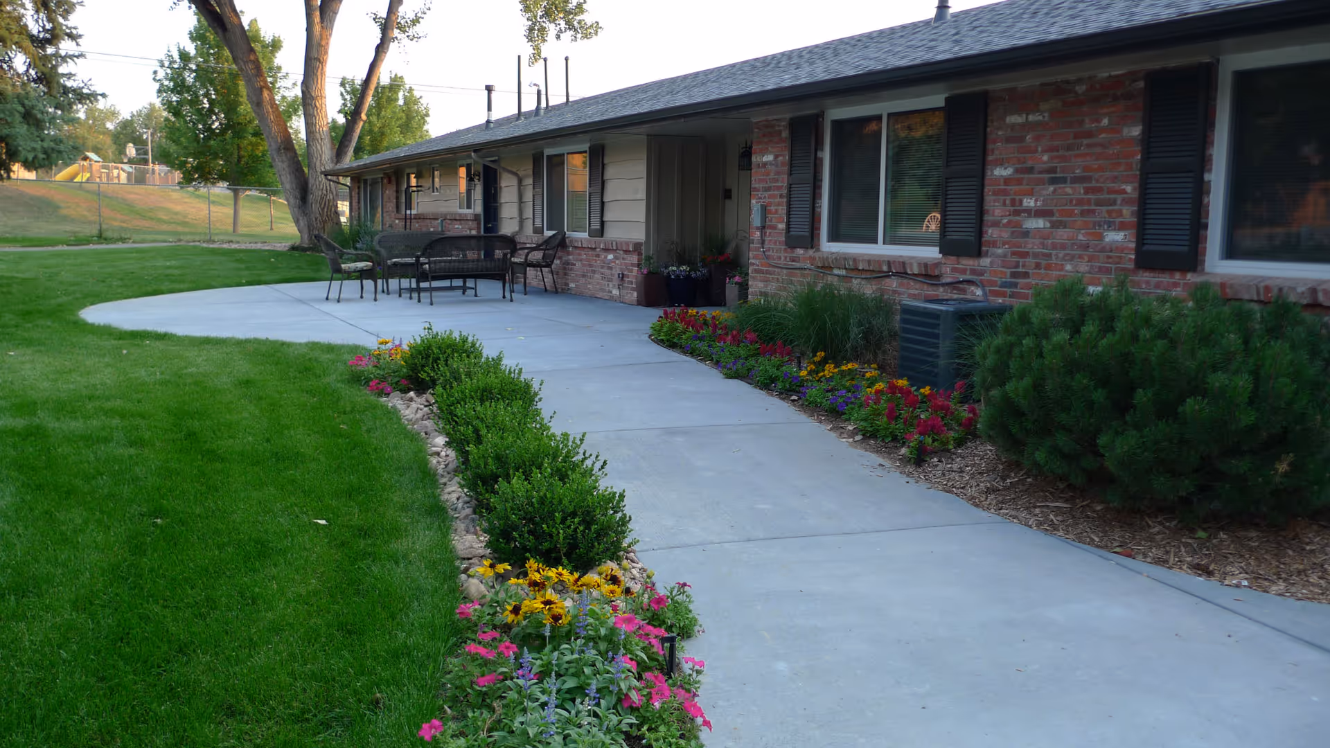 A paved walkway leading to the entrance of a single-story brick building with black shutters. The walkway is bordered by green grass and colorful flower beds with various shrubs. There is outdoor patio furniture including a table and chairs near the entrance. Trees and a playground are visible in the background.