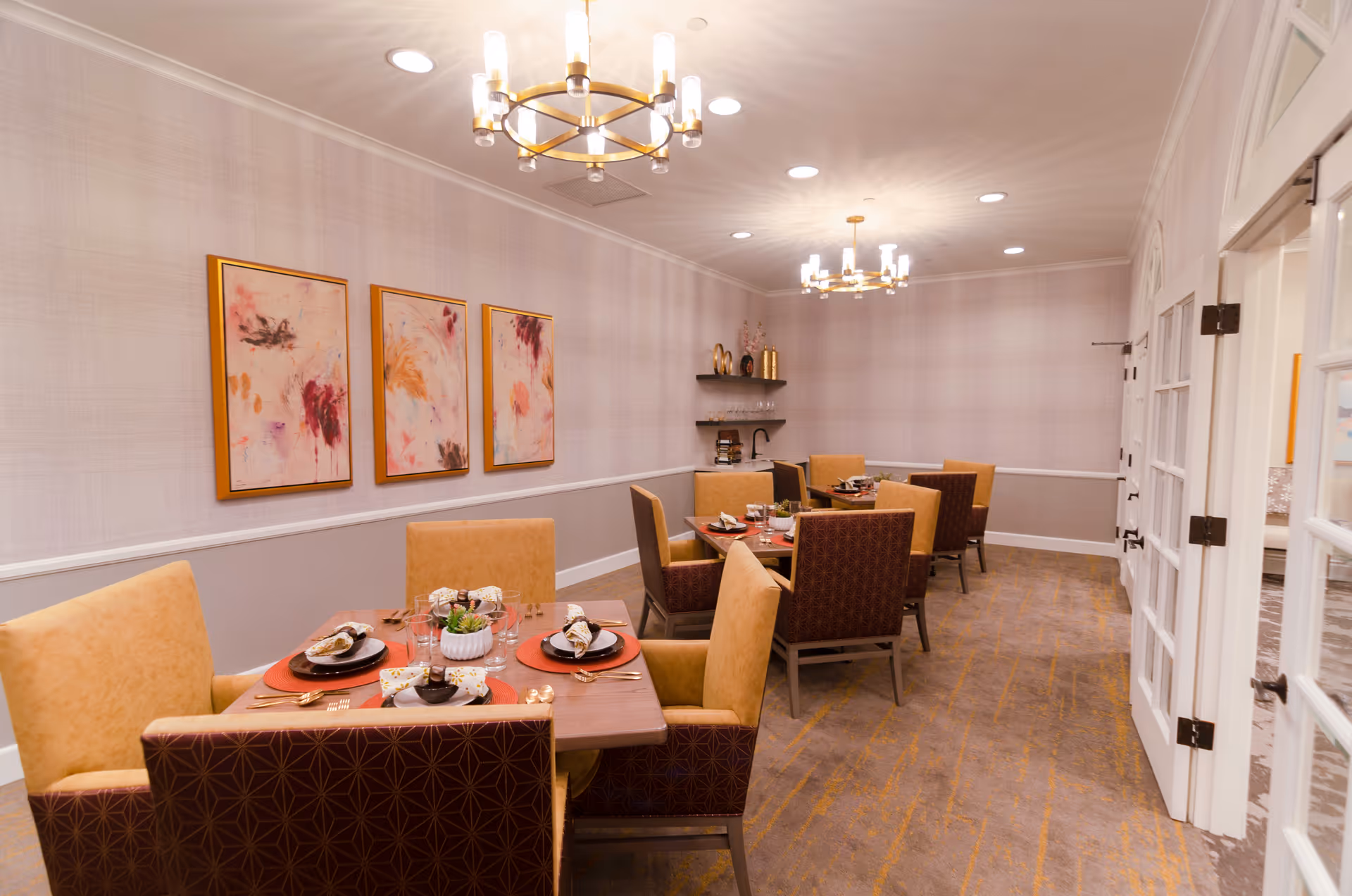 Well-lit dining room with several set tables, upholstered chairs, chandeliers, and abstract artwork on the wall.