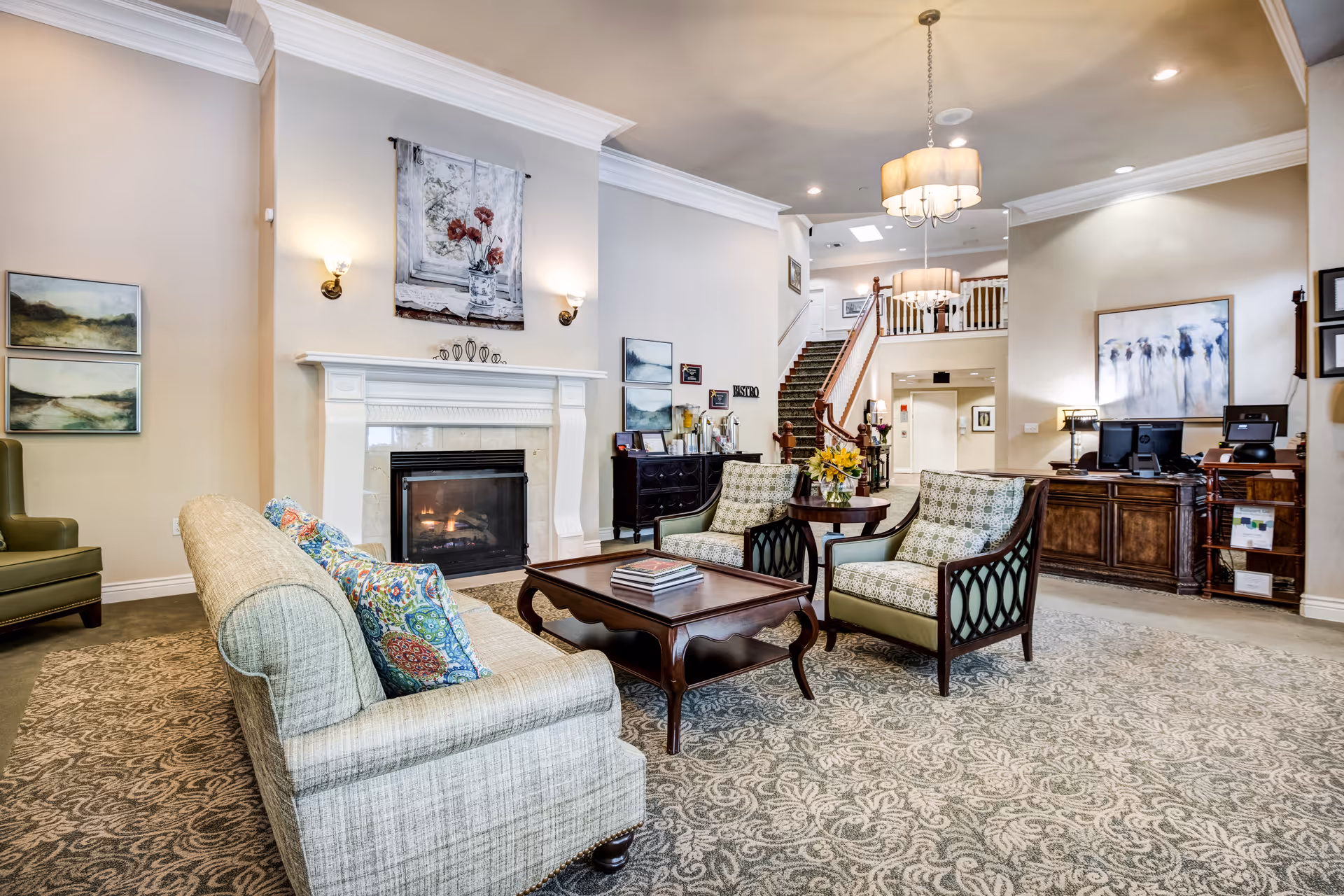 A spacious and elegant living room area in a senior living facility featuring a beige sofa with colorful pillows, two patterned armchairs, a wooden coffee table with books, a fireplace with a floral painting above it, and a staircase leading to an upper floor. The room is decorated with wall art, a chandelier, and has a reception desk with computer monitors in the background.