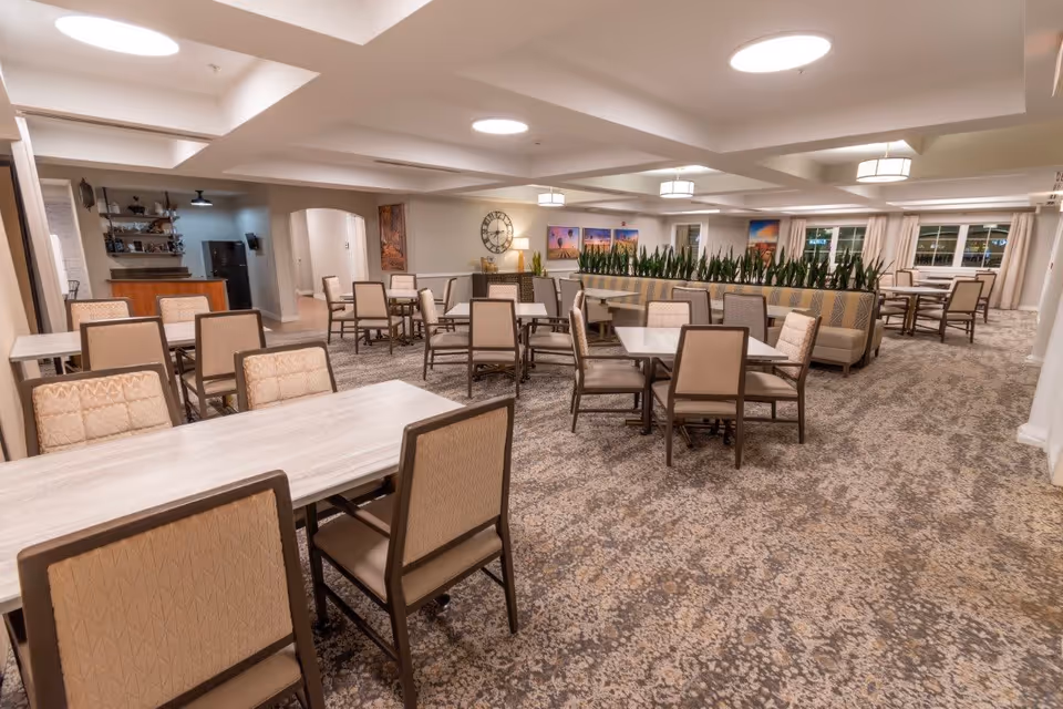 A spacious dining room with multiple tables and chairs arranged neatly. The room features a patterned carpet, ceiling lights, and a divider with tall green plants. There are framed pictures and a large clock on the walls, and windows with curtains letting in natural light.