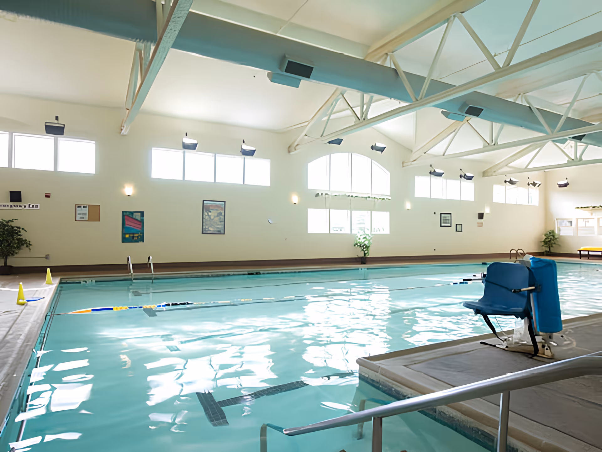 Indoor swimming pool with clear water, lane dividers, and a pool lift chair for accessibility. The pool area has large windows letting in natural light, white walls with framed artwork, and a high ceiling with exposed beams.