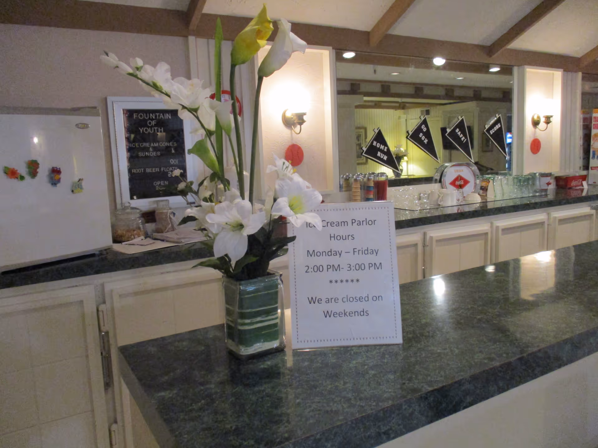 Interior view of an ice cream parlor counter with a vase of white flowers and a sign displaying the ice cream parlor hours. Behind the counter are various ice cream cones, cups, and a mirror reflecting pennants with phrases like 'HOME RUN' and 'GO BOX'.