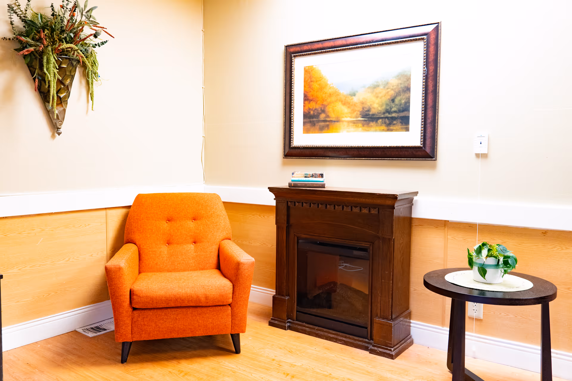 A cozy corner in a room featuring an orange upholstered armchair, a dark wooden electric fireplace, a round side table with a small potted plant, a framed landscape painting on the wall, and a decorative wall-mounted plant arrangement.