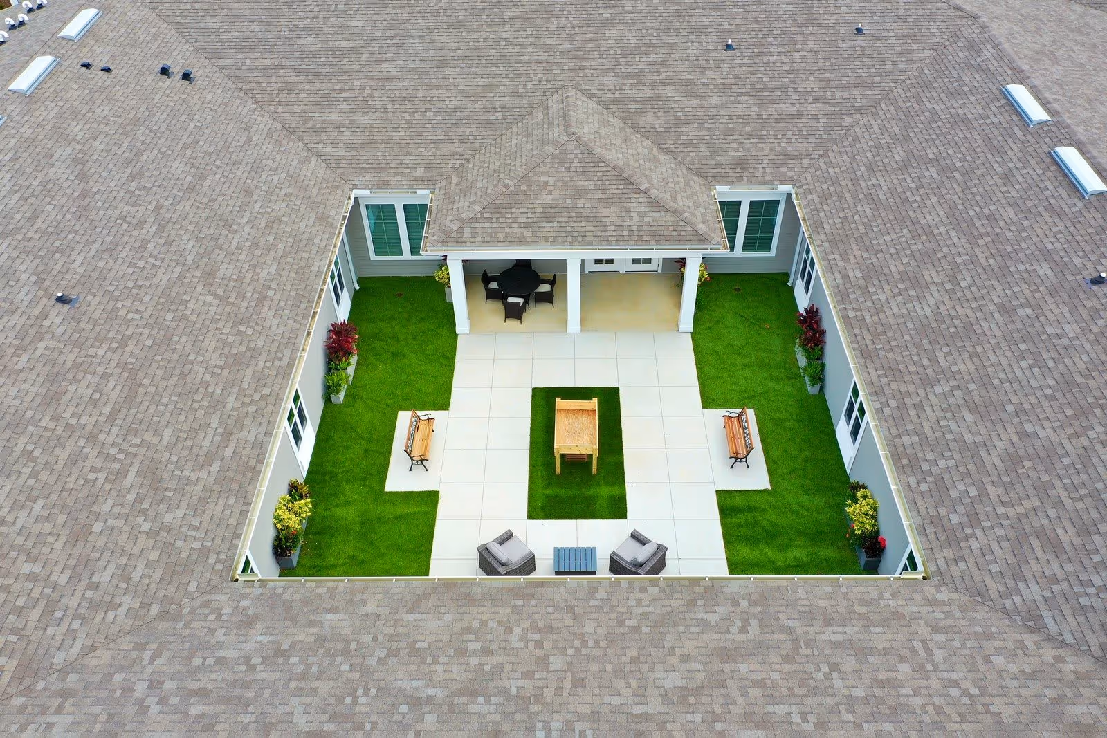 Aerial view of a courtyard enclosed by a building roof, featuring a central paved area with a wooden table, surrounded by green artificial grass, two benches on either side, two armchairs with a small table, potted plants, and a covered patio area with a round table and chairs.