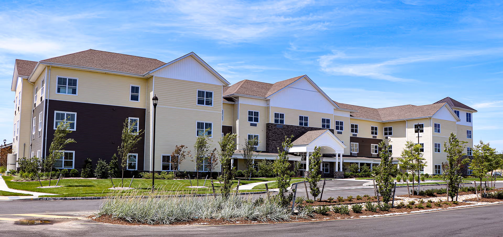 Exterior view of a large, modern three-story senior living facility building with beige and brown siding, multiple windows, a covered entrance, and landscaped grounds with young trees and shrubs under a blue sky.