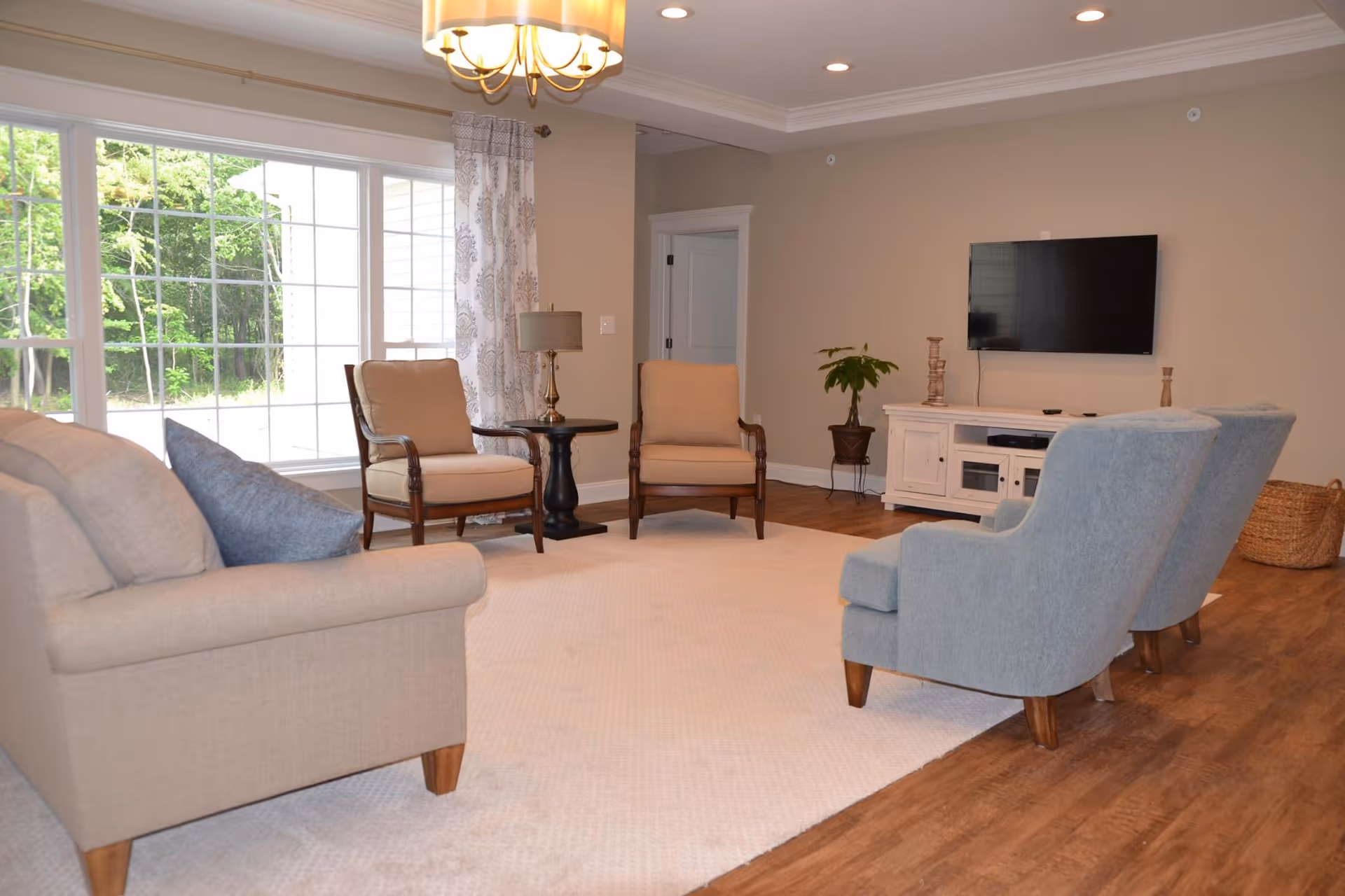 A cozy living room with beige and blue upholstered chairs and a beige sofa arranged around a large cream-colored rug. There is a large window with patterned curtains letting in natural light, a small round side table with a lamp, a white media console with a flat-screen TV mounted on the wall above it, and a potted plant in the corner. The room has wooden flooring and beige walls.