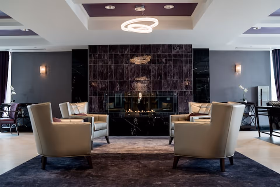 A modern living room with four beige armchairs arranged around a dark purple rug facing a sleek black fireplace with a tiled wall. The room features dark walls with wall sconces, side tables with white orchids, and a contemporary circular ceiling light fixture.