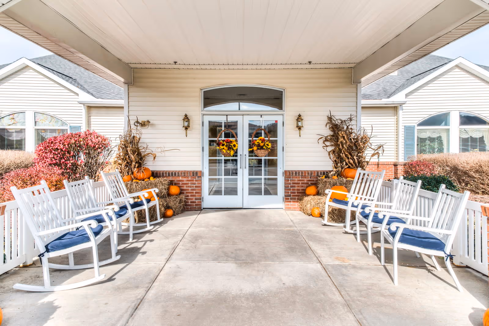 Entrance of Walden Place facility with double glass doors decorated with autumn wreaths. The porch area features white rocking chairs with blue cushions on both sides, surrounded by fall decorations including pumpkins, hay bales, and dried corn stalks. The building exterior is light-colored with brick accents and windows with white trim.