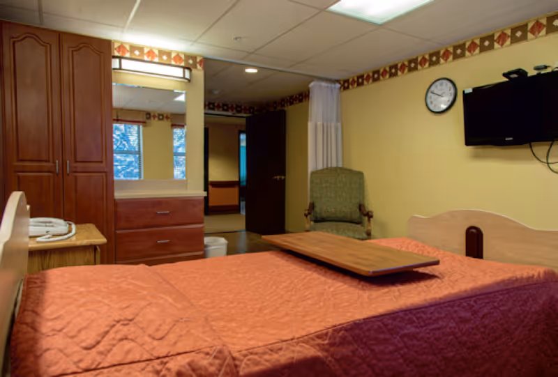 A nursing facility private bedroom with a bed covered by a red quilt, wall-mounted TV, dresser, wardrobe and an armchair.