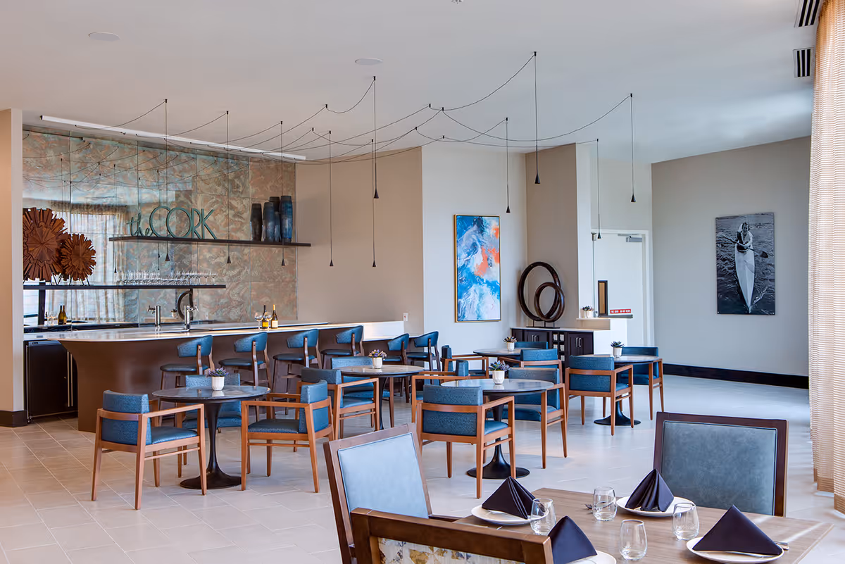 A modern dining area with multiple round tables and blue cushioned chairs. In the background, there is a bar counter with high stools and decorative items on shelves. The walls feature abstract artwork and a black-and-white photo of a person in a canoe. Pendant lights hang from the ceiling, and the room has large windows with sheer curtains allowing natural light to enter.