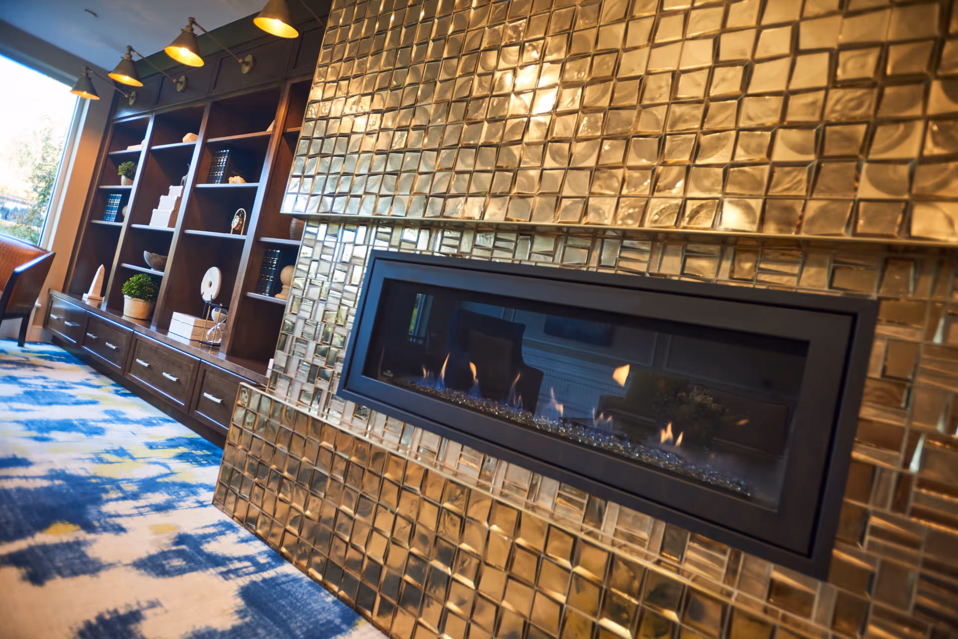 A modern interior space featuring a long, rectangular gas fireplace set into a wall covered with shiny, gold-colored square tiles. To the left, there is a built-in dark wood shelving unit with various decorative items and books, illuminated by wall-mounted lights above. The floor has a patterned carpet with blue, yellow, and white colors.