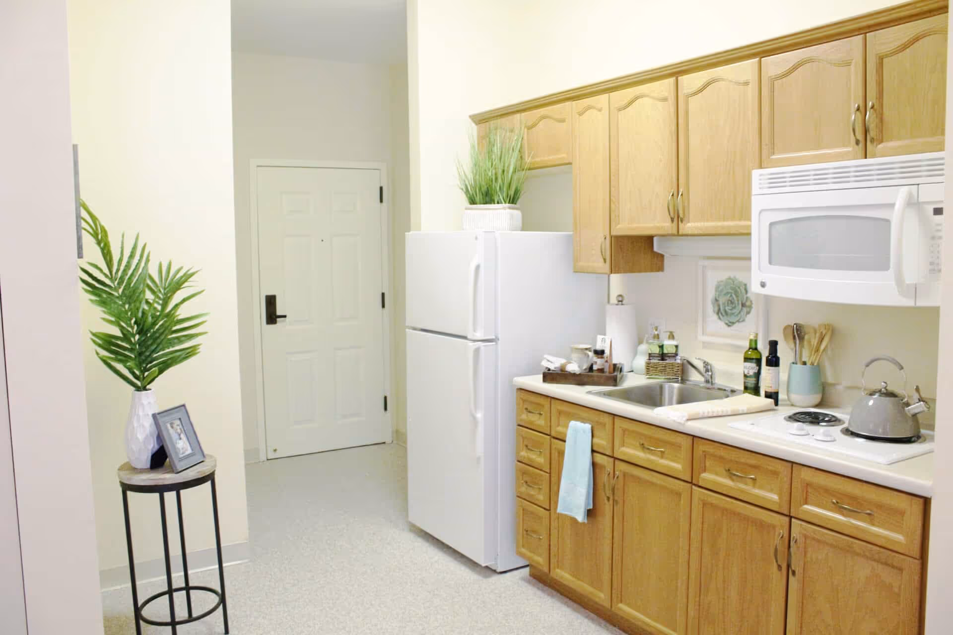 A small kitchen area with wooden cabinets, a white refrigerator, a microwave, a sink, and a stovetop with a kettle. There is a small round table with a white vase holding green plants and a framed photo. The walls are light-colored and there is a door visible in the background.