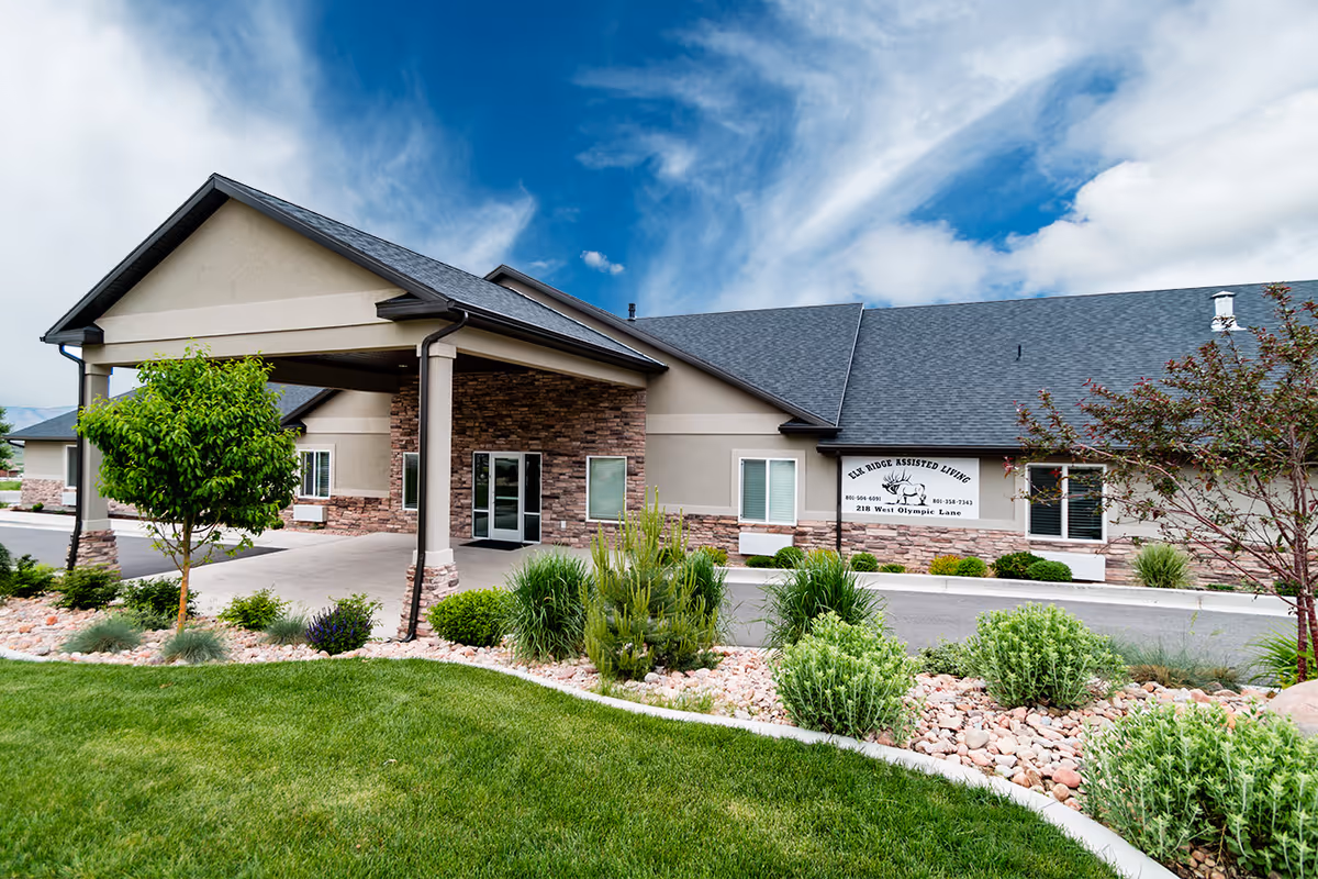 Exterior view of Elk Ridge Assisted Living facility showing a single-story building with a covered entrance, stone and beige siding, surrounded by well-maintained landscaping including green grass, bushes, and small trees under a partly cloudy blue sky.