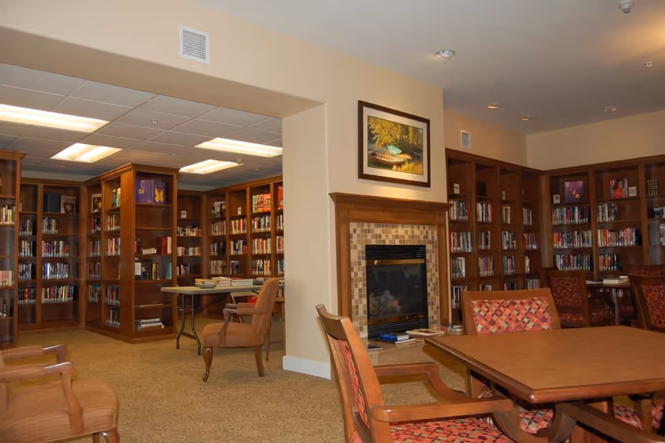 Interior of a senior living facility library or reading room with wooden bookshelves filled with books, a fireplace with a framed painting above it, several wooden tables and chairs with patterned upholstery, and a carpeted floor.