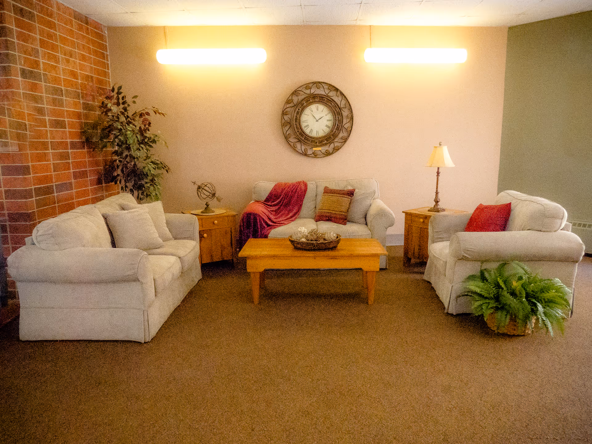 A cozy living room area with three beige upholstered sofas arranged around a wooden coffee table. The coffee table has a decorative basket with natural elements. Behind the middle sofa, there is a round wall clock and two wall-mounted lights. On either side of the middle sofa are wooden side tables, one with a small globe and the other with a lamp. There is a potted plant on the left side near a brick wall and a fern plant on the right side near the armchair.