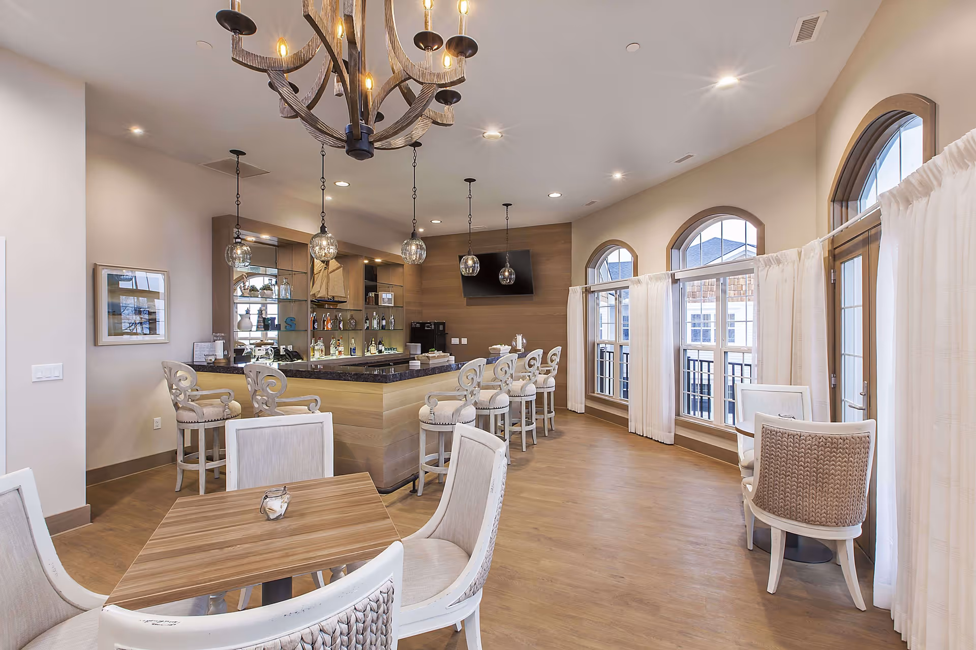 A bright and spacious interior room featuring a bar area with high stools, a countertop with bottles and glassware, and a mounted TV. The room has large arched windows with white curtains allowing natural light to fill the space. There are several small tables with cushioned chairs arranged around the room, and elegant pendant lights and a chandelier hanging from the ceiling.