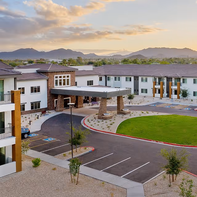 Exterior view of ACOYA Mesa senior living facility at sunset, showing a large building with multiple windows, a covered entrance supported by stone pillars, a circular driveway, parking spaces including handicapped spots, landscaped areas with small trees and bushes, and mountains in the background under a partly cloudy sky.