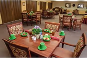 Dining room in a senior living facility with wooden tables set with green cups and plates, floral centerpieces, and upholstered chairs.