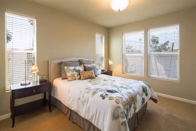 A bright bedroom with a bed featuring floral bedding and multiple pillows. There are two dark wooden nightstands on either side of the bed, each with a lamp. The room has beige walls and carpet, and three large windows with white blinds letting in natural light.