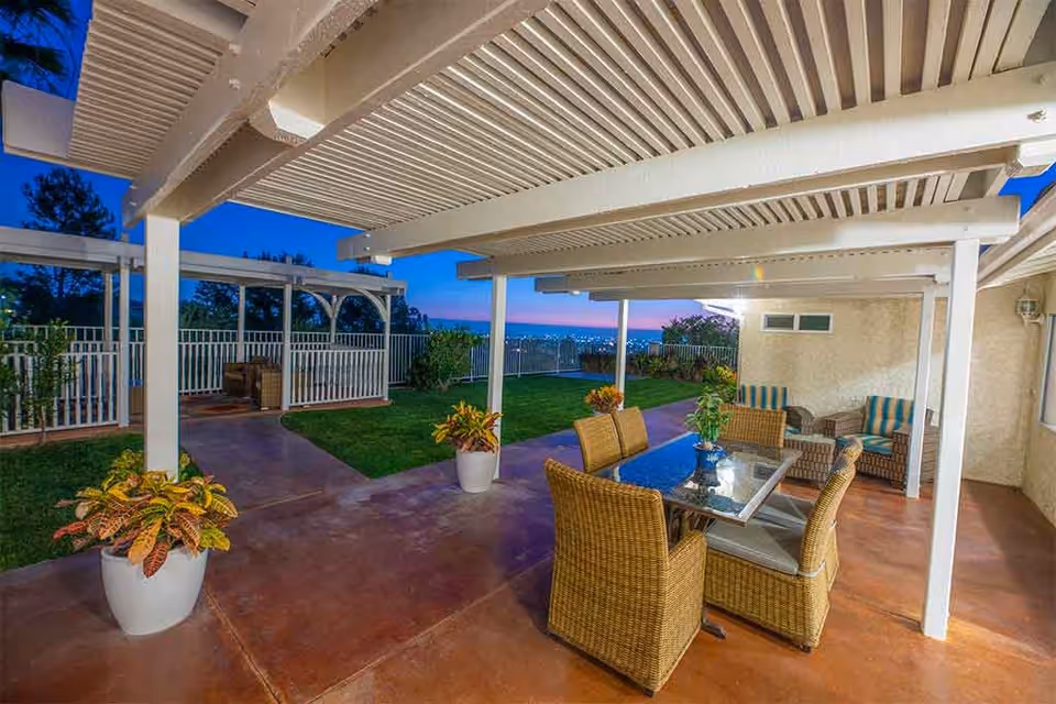 Outdoor patio area at dusk with a white pergola overhead, a glass-top table surrounded by wicker chairs, potted plants, and cushioned wicker seating against the wall. The area overlooks a fenced garden with a view of city lights in the distance.