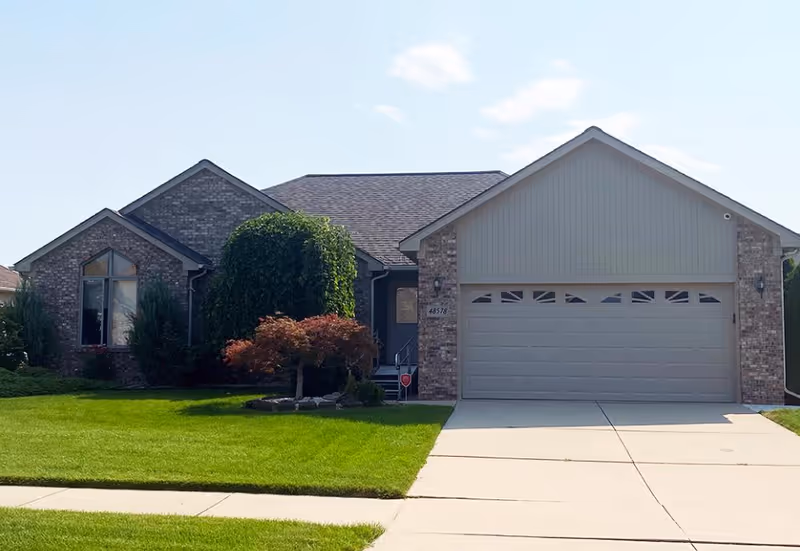 Front exterior view of a single-story brick house with a two-car garage, well-maintained green lawn, and small trees and shrubs in the yard under a clear sky.