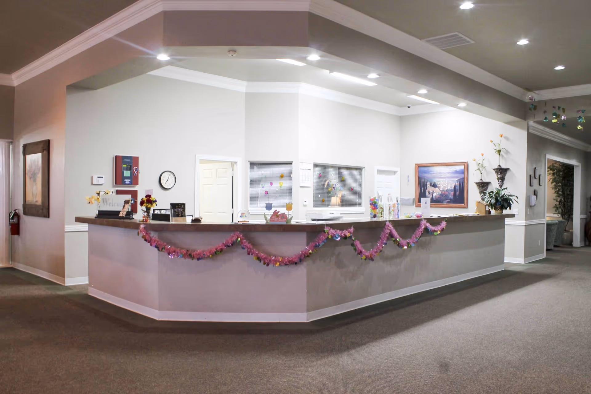 Reception desk area in a senior living facility decorated with pink garlands and various small decorations. The desk has a wooden countertop with flowers, a clock on the wall, and windows with blinds behind it. The area is well-lit with ceiling lights and has neutral-colored walls and carpeted flooring.