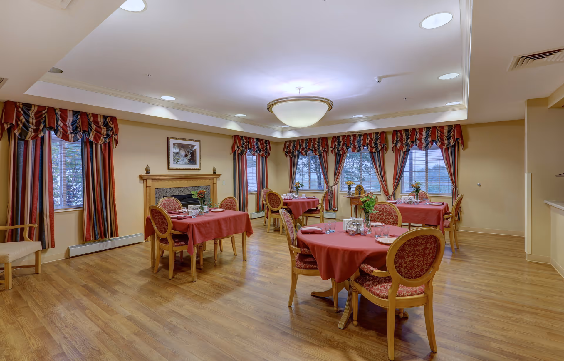 A dining room with several round and square tables covered with red tablecloths, each set with plates, glasses, and floral centerpieces. The room has wooden flooring, large windows with striped red, white, and blue curtains, and a ceiling light fixture. There is a fireplace with a framed picture above it on one wall.