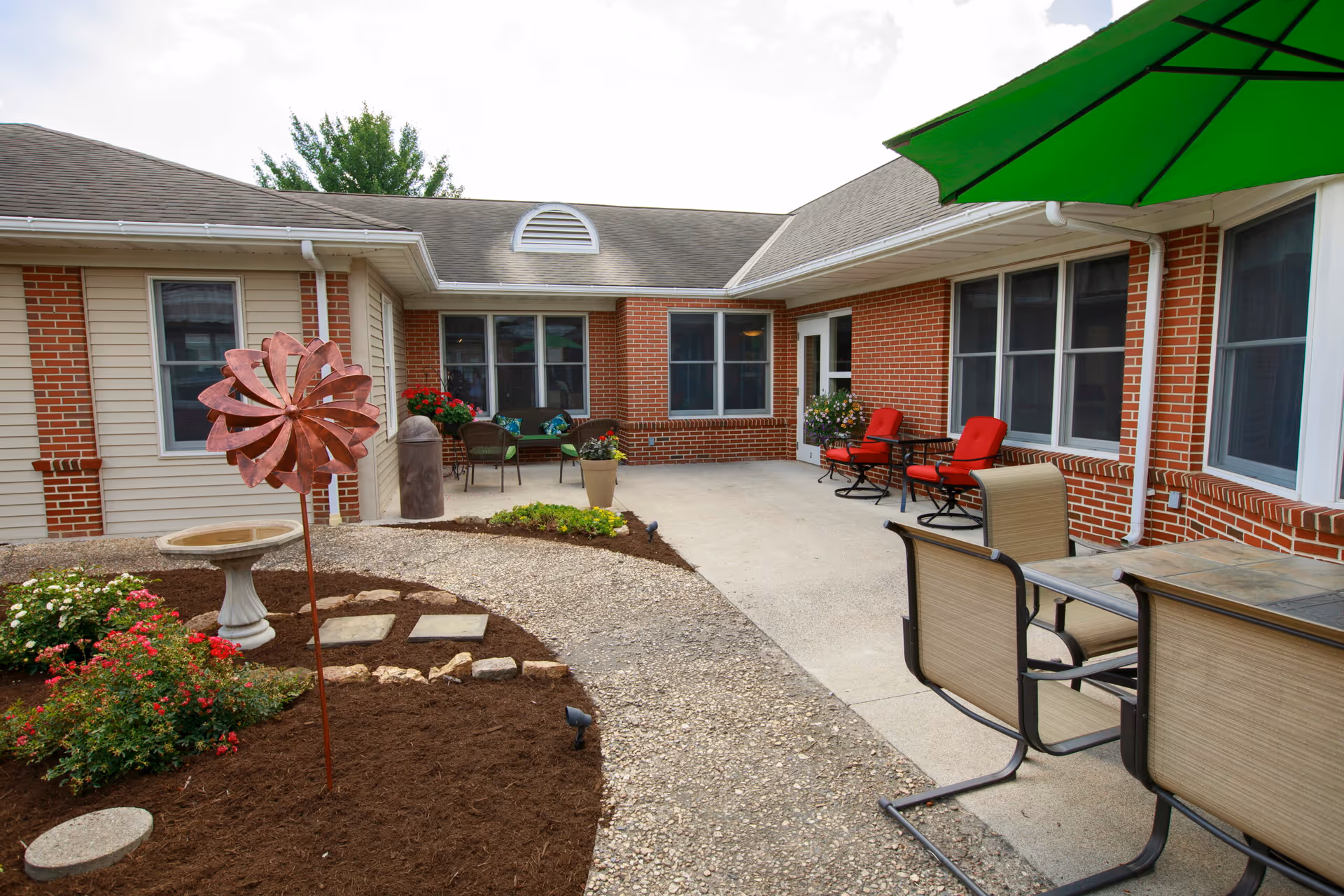 Outdoor patio area at Valley View Retirement Community featuring a paved walkway, garden with flowers and a birdbath, patio furniture including chairs and tables, and a large green umbrella. The building exterior is visible with brick and siding walls and multiple windows.