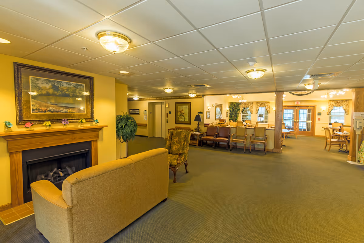 A spacious senior living facility common area with a beige couch and an upholstered armchair facing a wooden fireplace with a framed landscape painting above it. The room has yellow walls, carpeted floors, and ceiling lights. In the background, there are several chairs and tables arranged near windows with decorative curtains, and an elevator is visible on the left side.