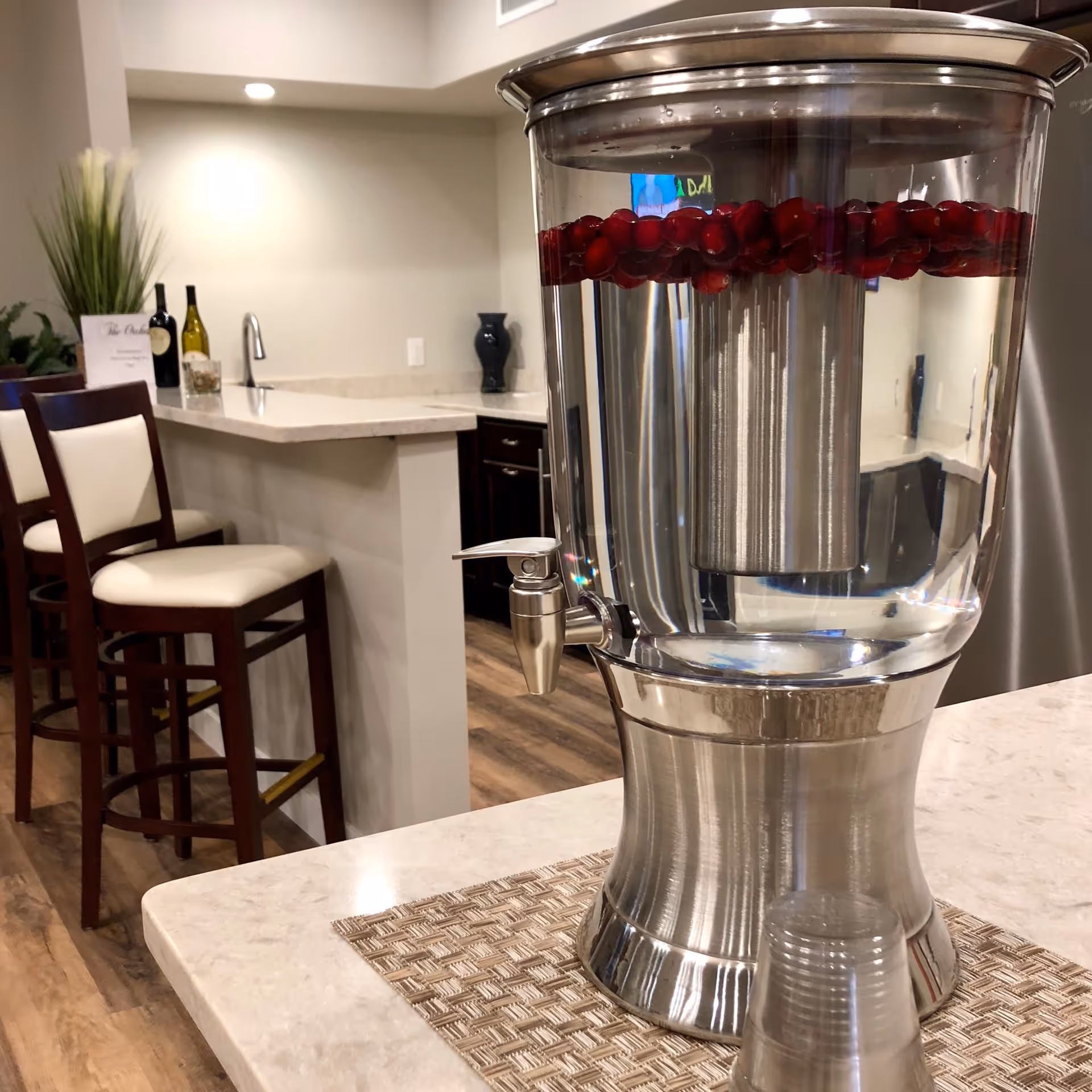 A modern kitchen or dining area with a large stainless steel water dispenser filled with water and floating cranberries on a countertop. In the background, there are two high chairs with white cushions, a countertop with a sink, two bottles of wine, a small plant, and a decorative vase.