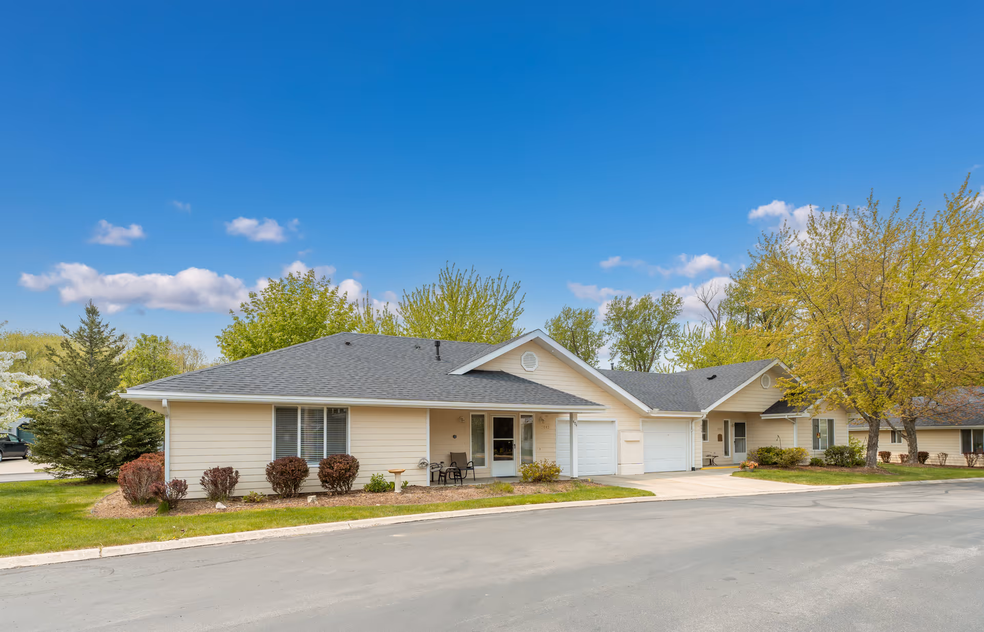 Exterior view of a single-story residential building with beige siding and a dark gray roof. The building has a small front porch with two chairs and a table, a garage, and is surrounded by green grass, bushes, and trees under a clear blue sky.