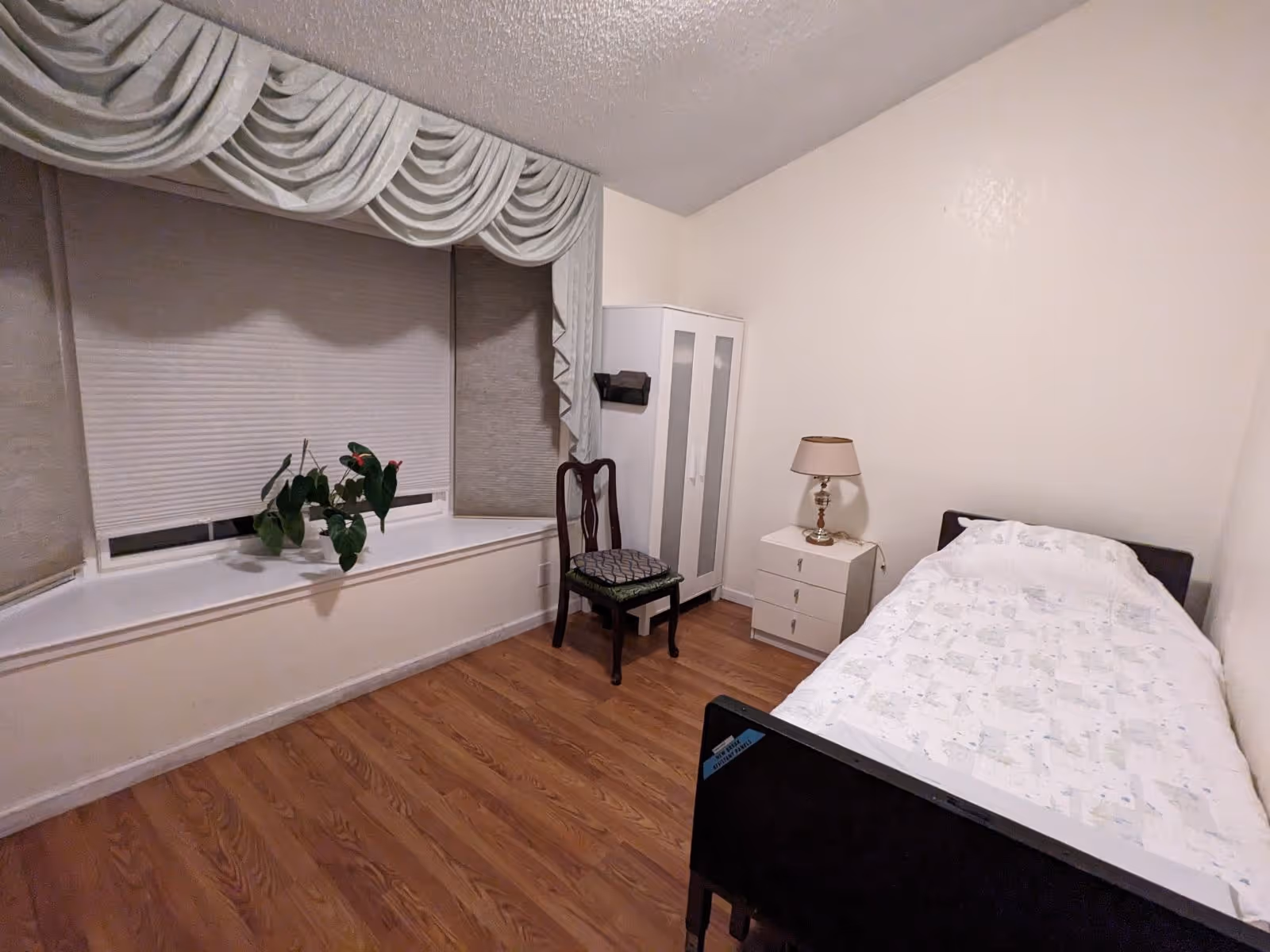 Simple bedroom with a single bed, nightstand and lamp, wardrobe and chair, and a window seat with a potted plant.