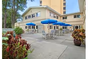 Outdoor patio area at Mease Life facility with multiple tables and chairs shaded by blue umbrellas, surrounded by potted plants and trees, adjacent to a multi-story building under a clear sky.