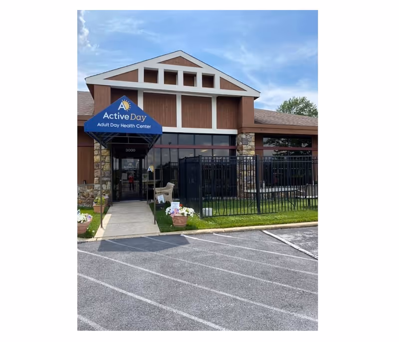 Front entrance of Active Day Adult Day Health Center showing a blue awning, glass doors, fenced patio and parking area.
