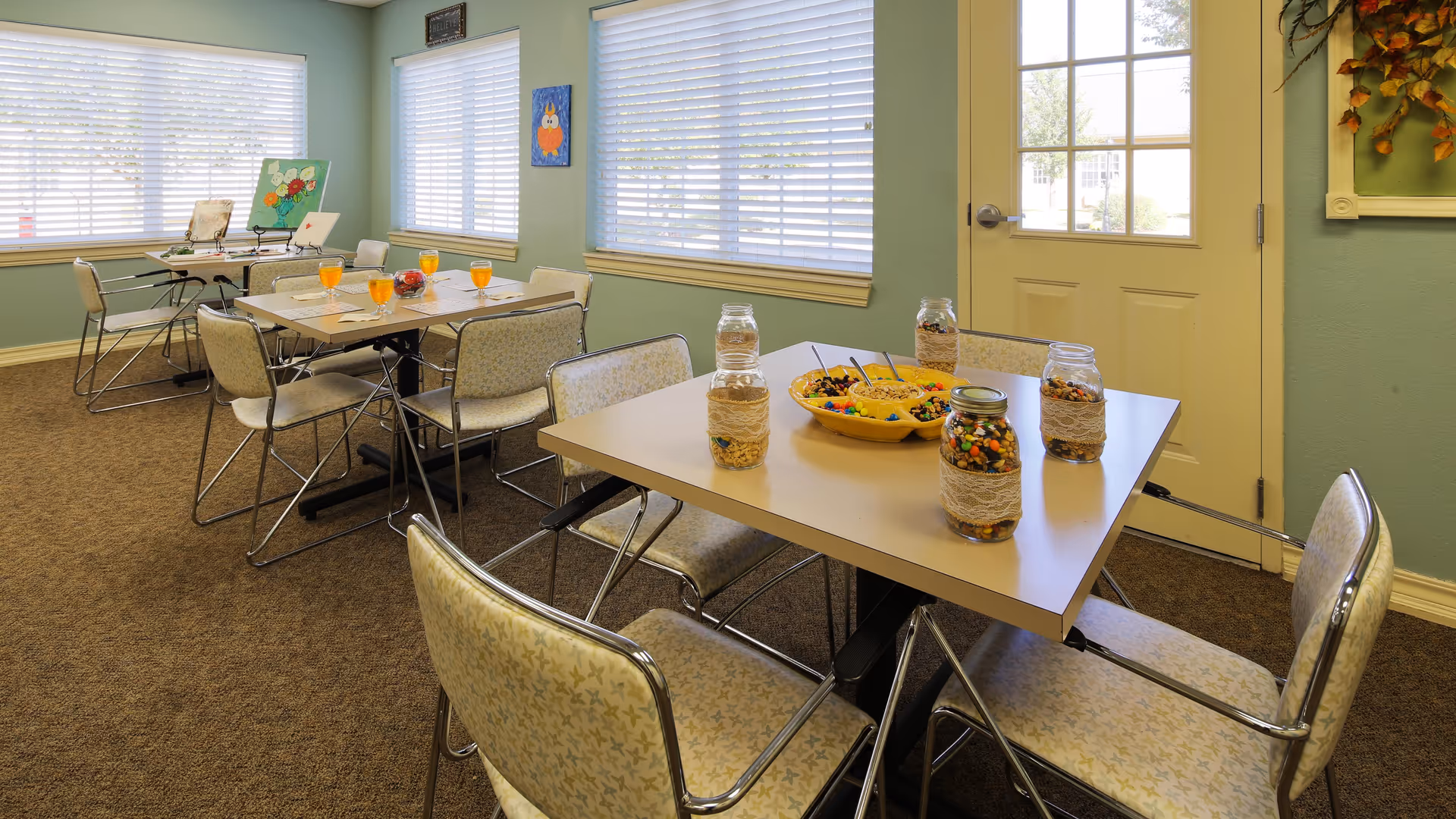 A bright room with multiple tables and chairs arranged for dining or activities. On the tables are jars filled with colorful candies and snacks, along with a bowl of mixed treats. The room has large windows with blinds, light green walls, and a door with glass panels. The carpet is brown, and there are a few pieces of wall art visible.